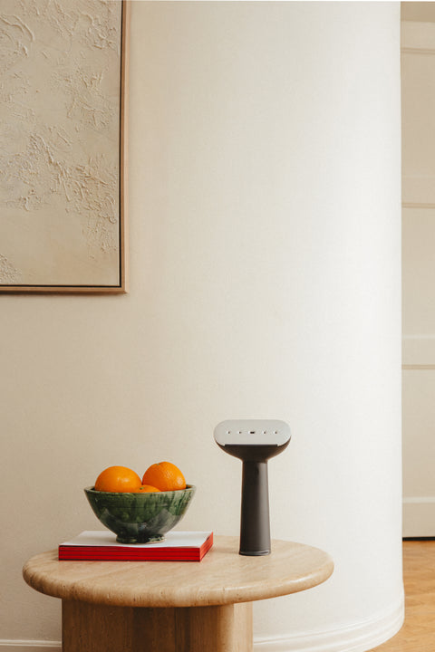 Modern interior with a small table holding a bowl of fruit and a handheld steamer, with a white wall and door in the background.