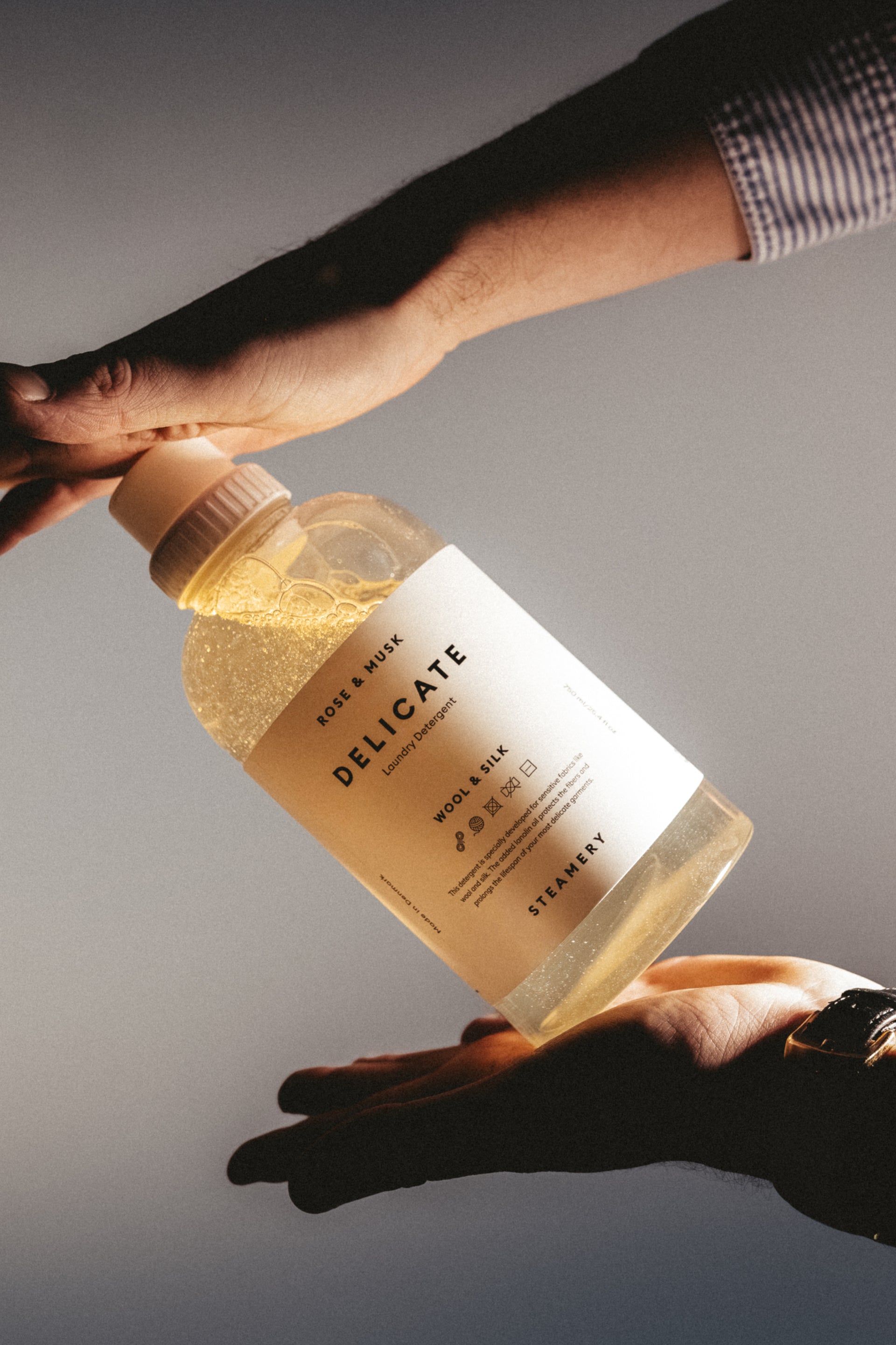 Bottle of 'Delicate' laundry detergent being passed between two hands on a neutral background