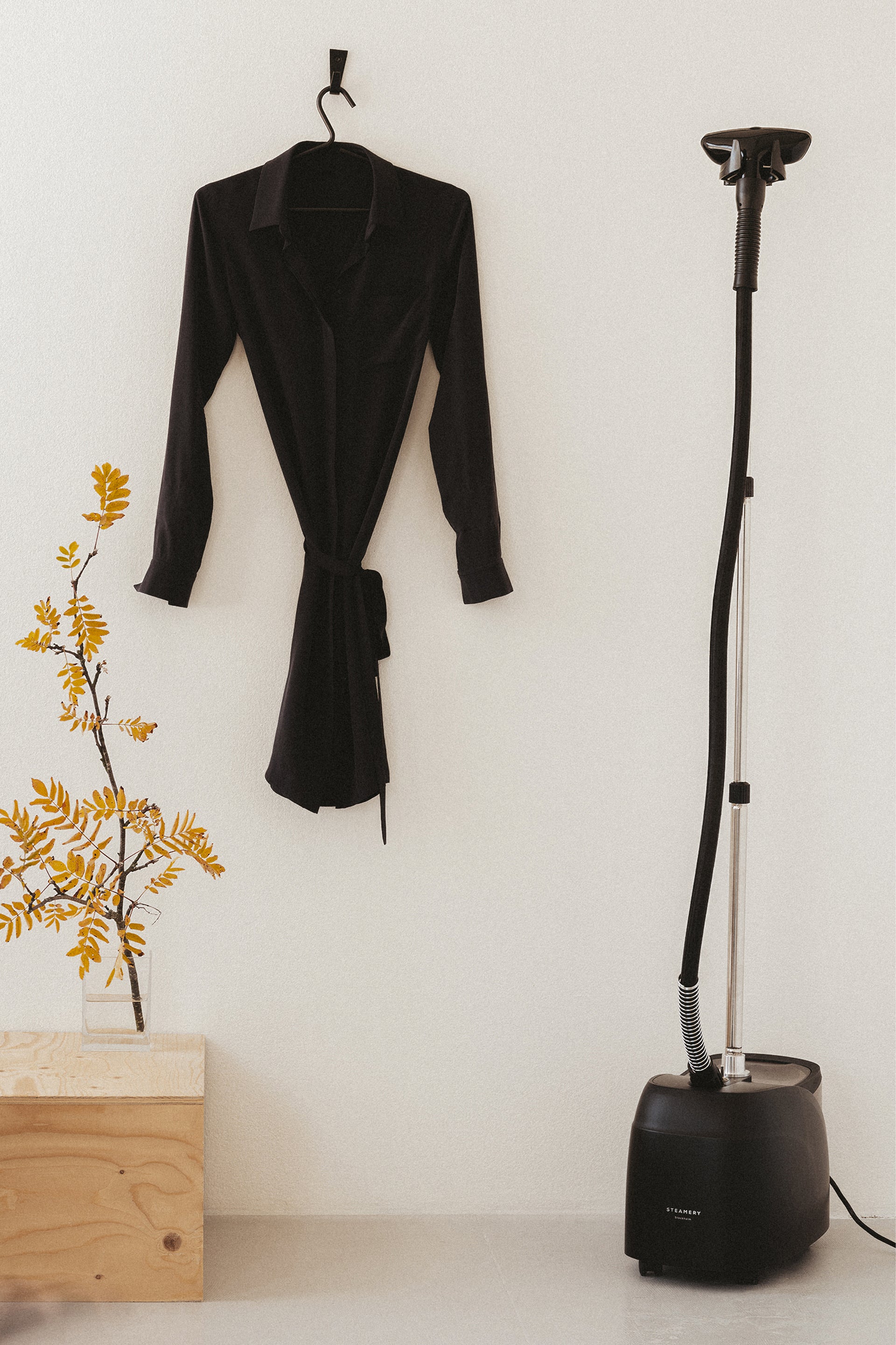 Black garment steamer next to a black dress on a hanger against a white wall.