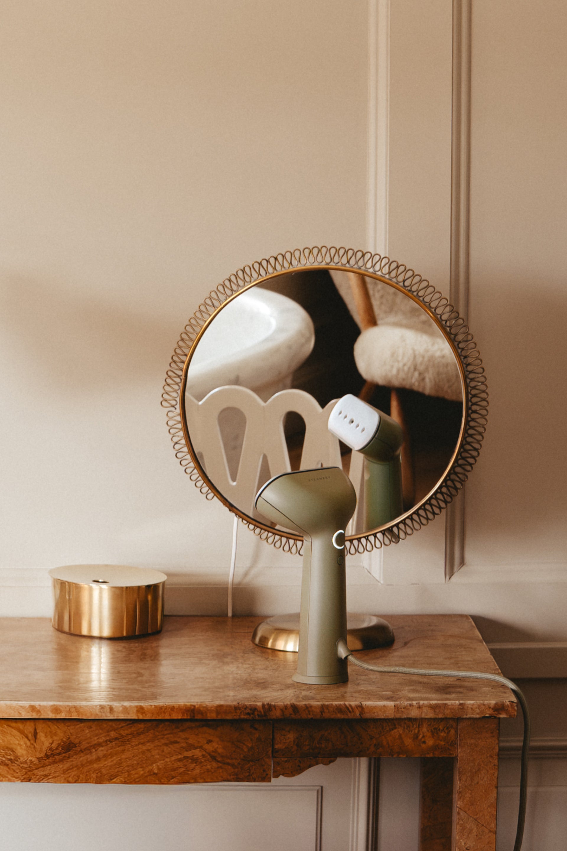 Green steamer in front of a round mirror with decorative frame on a wooden surface against a beige wall.