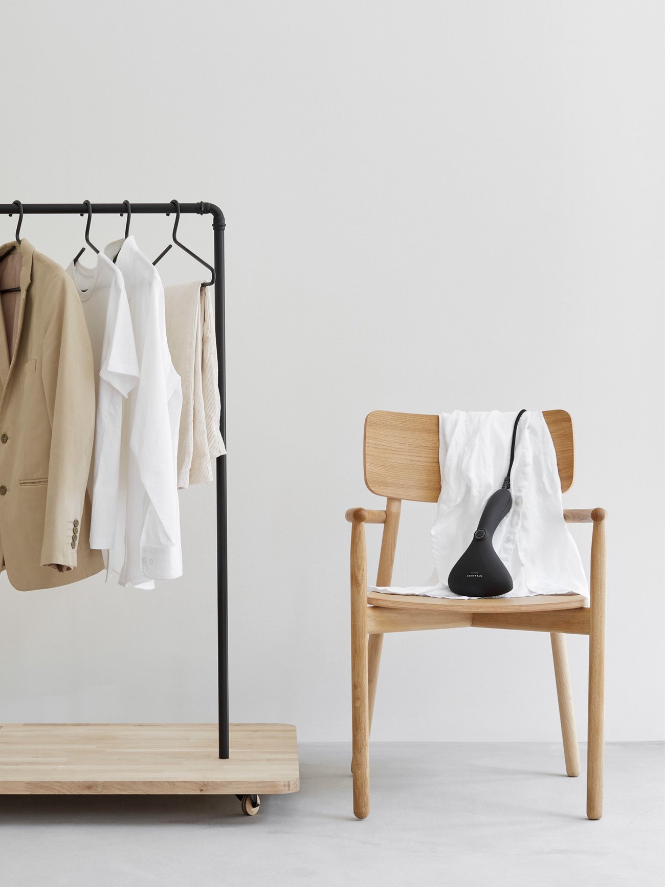 The Steamery Cirrus 2 Handheld Steamer rests over a chair beside a clothing rack.